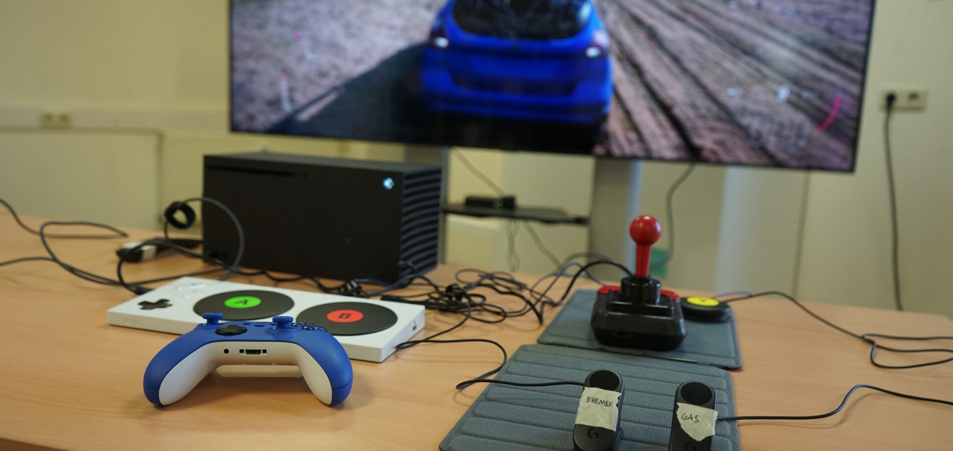 Eine totale Ansicht zeigt verschiedene Gamingsetups. Im Hintergrund ist ein TV-Bildschirm mit einem Autorennen zu sehen. Auf einem Tisch davor sind verschiedene Controller zu sehen.