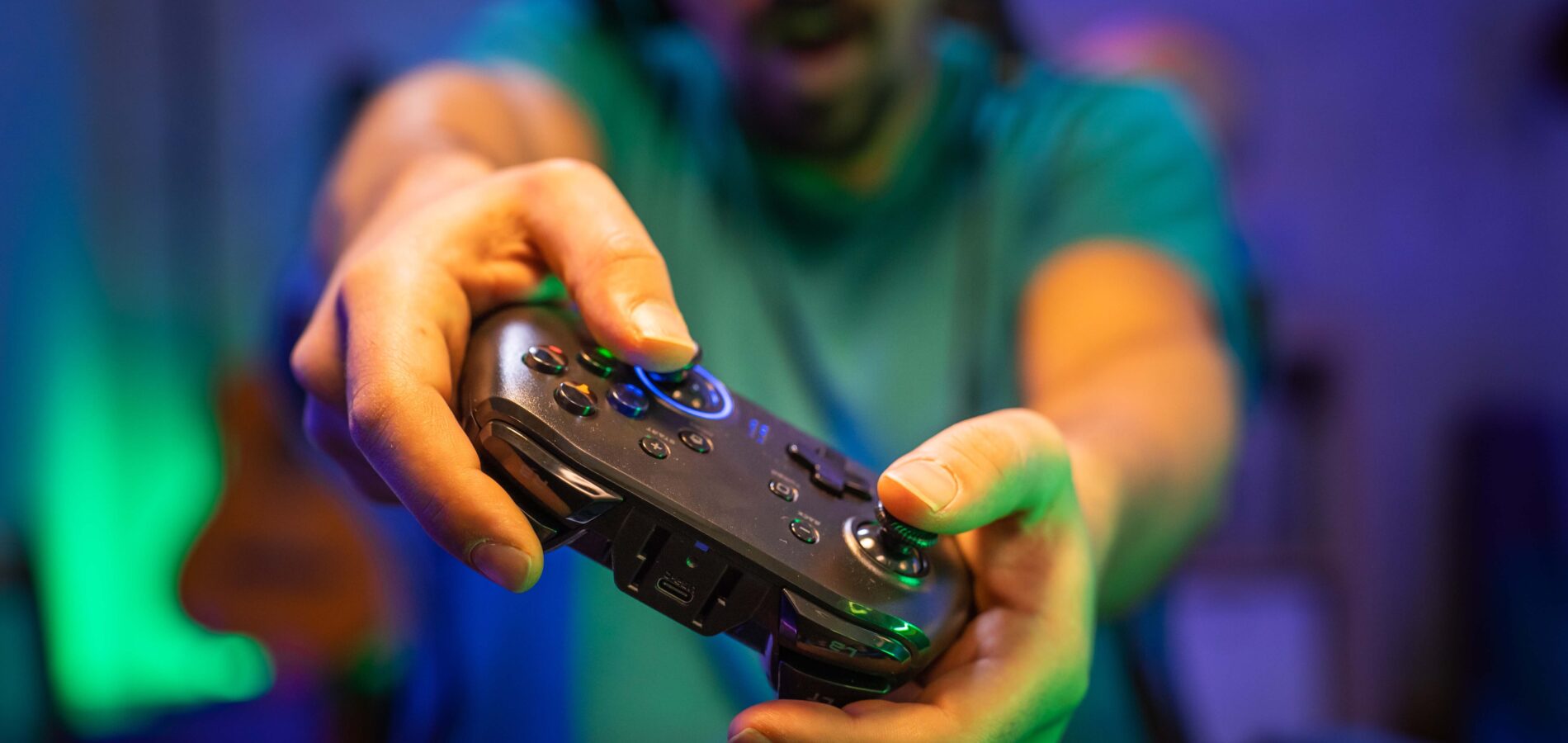 Im Fokus ist ein schwarzer Gamingcontroller, gehalten von einer männlichen Personen. Das Bild wurde während des Spiels gemacht. Der Mann drückt verschiedene Knöpfe und neigt den Controller.