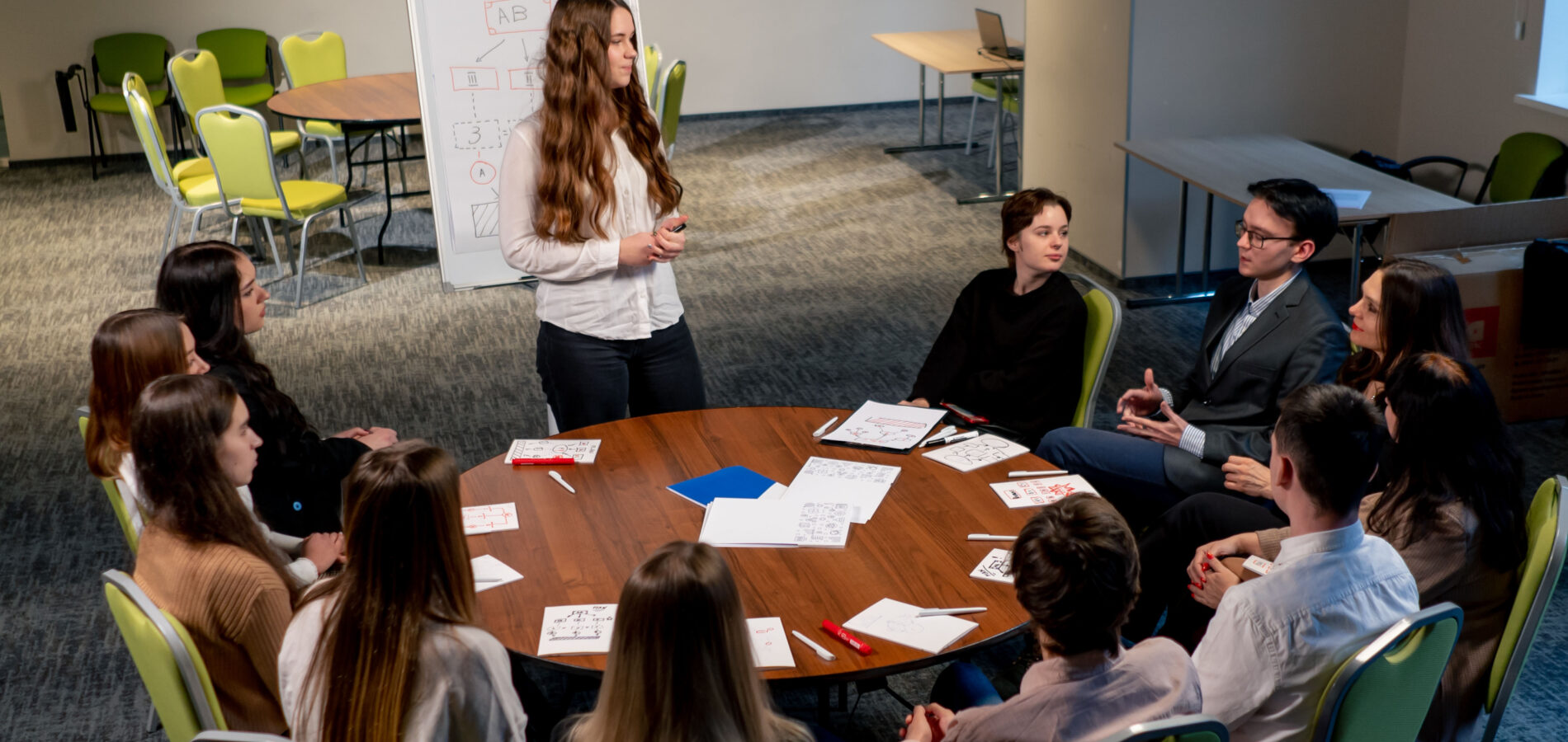 Zehn Personen sitzen in einem Konferenzraum an einem runden Tisch. Eine weitere Person steht am Tisch und moderiert das Gespräch zwischen den anderen Personen.