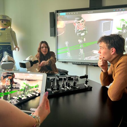 Ein großer runder Tisch steht vor einem TV-Bildschirm. Daran sitzen drei Personen. Zwei davon unterhalten sich miteinander, eine dritte Person hält ein Tablet in der Hand. Auf dem Tisch befinden sich VR-Brillen unterverschiedene weitere VR-Utensilien.