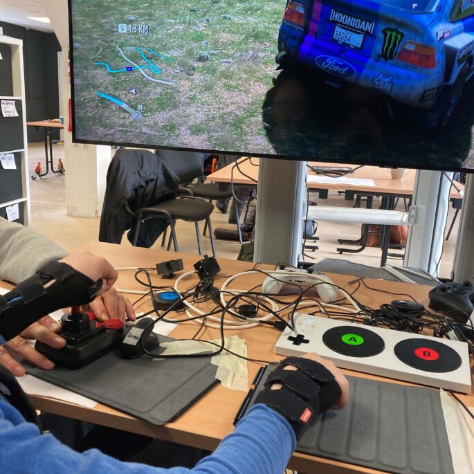 Ein Gamingsetup wird von einer Person bedient. Im Hintergrund ist ein TV-Bildschirm mit einem Autorennen zu sehen. Auf einem Tisch davor sind verschiedene Controller zu sehen.