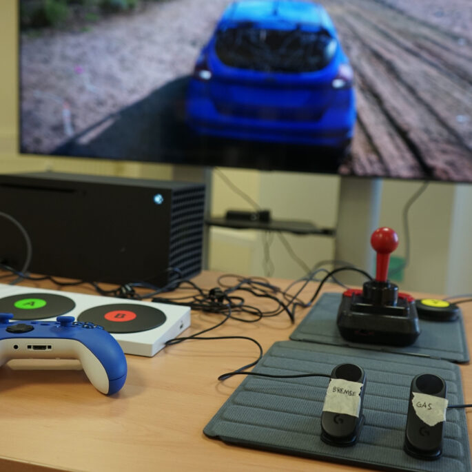 Eine totale Ansicht zeigt verschiedene Gamingsetups. Im Hintergrund ist ein TV-Bildschirm mit einem Autorennen zu sehen. Auf einem Tisch davor sind verschiedene Controller zu sehen.