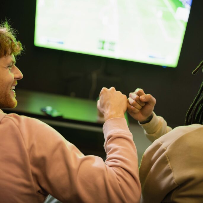 Zwei Männer sitzen vor einem Bildschirm und sind von hinten zu sehen. Einer schaut den anderen an. Im gleichen Moment geben sie sich eine Faust und strahlen Freude und Spaß aus.