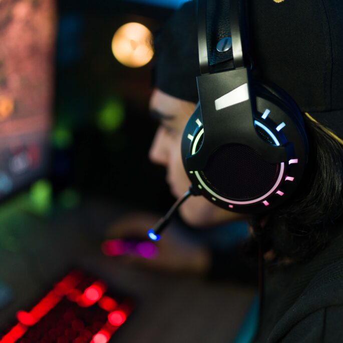 Ein Gamer sitzt vor einem PC-Bildschirm an einem Schreibtisch. Vor ihm liegen eine Gaming-Tastatur und eine Gaming-Maus. In der Seitansicht ist zu erkennen, dass er eine Cap und ein Gaming-Headset trägt. Er ist vertieft in das Spiel.