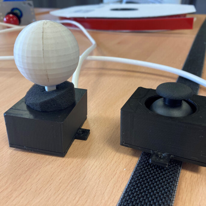 Zwei barrierefreie Joysticks stehen auf einem Tisch nebeneinander. Einer davon in auf einem Klettband befestigt, damit der Joystick nicht verrutscht.