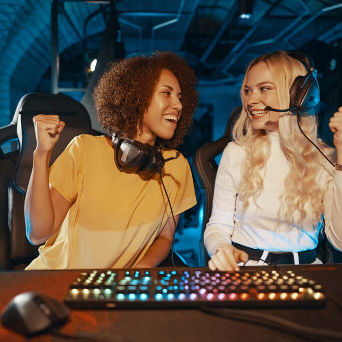 Zwei junge Frauen sitzen an einem Schreibtisch. Vor ihnen liegt eine bunt leuchtende Gamingtastatur von einem PC. Beide Frauen schauen sich gegenseitig an und heben vor Freude Fäuste nach oben.