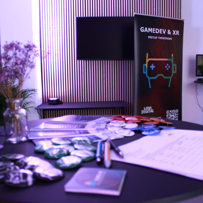 Im Vordergrund steht ein Stehtisch mit Werbematerialien. Diese ist unscharf im Bild. Der Fokus des Bildes liegt auf der Wand dahinter mit einem TV-Bildschirm und einem Roll-Up-Banner mit der Aufschrift "GameDev & XR Meetup Thüringen".