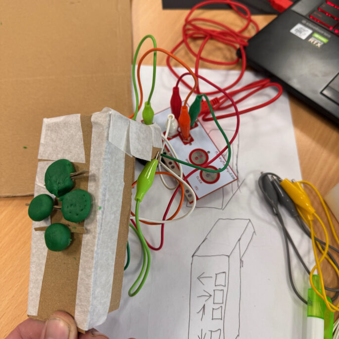 Auf einem Tisch liegen viele bunte Kabel, welche zur Programmierung von Mini-Computern genutzt werden. Als Unterlage dient ein selbst gezeichneter Bauplan. Die Kabel verbinden einen Laptop mit verschiedenen Elementen, die einen selbstgebauten Controller darstellen.