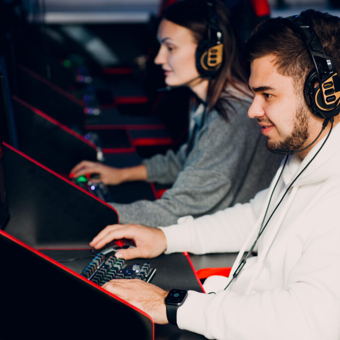 Zwei Personen sitzen vor Gaming-PCs in extra dafür angefertigten Stühlen. Diese Stühle erinnern an Sportwagensitze. Beide tragen Headsets und sind einem Spiel vertieft.