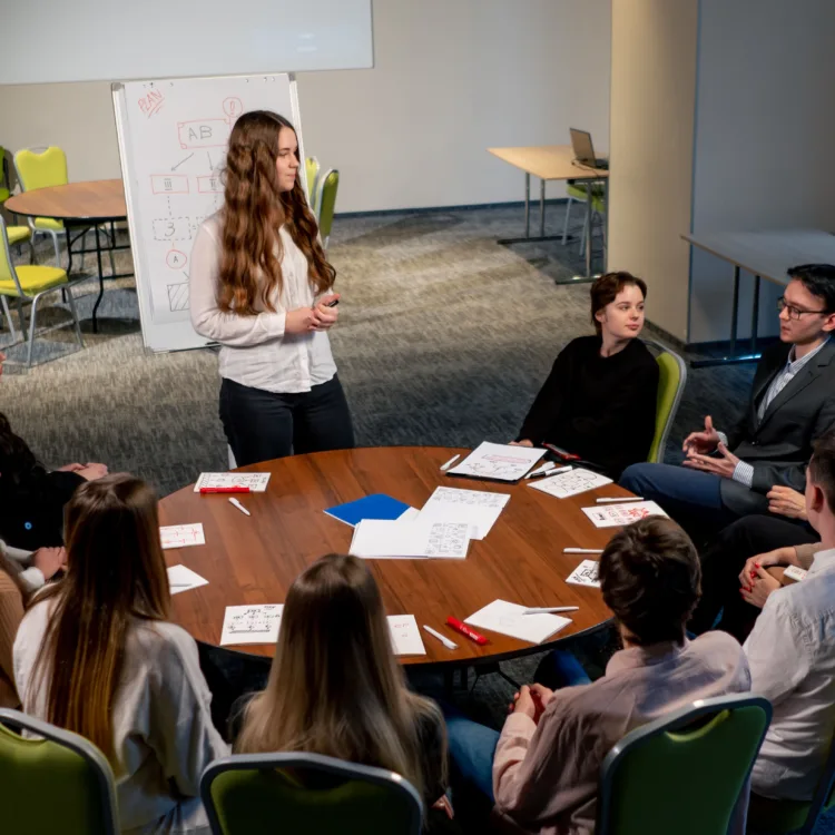 Zehn Personen sitzen in einem Konferenzraum an einem runden Tisch. Eine weitere Person steht am Tisch und moderiert das Gespräch zwischen den anderen Personen.
