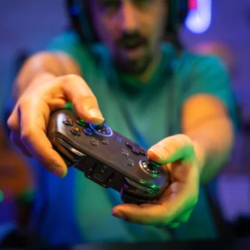 Im Fokus ist ein schwarzer Gamingcontroller, gehalten von einer männlichen Personen. Das Bild wurde während des Spiels gemacht. Der Mann drückt verschiedene Knöpfe und neigt den Controller.
