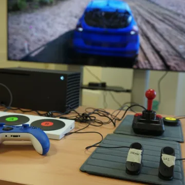 Eine totale Ansicht zeigt verschiedene Gamingsetups. Im Hintergrund ist ein TV-Bildschirm mit einem Autorennen zu sehen. Auf einem Tisch davor sind verschiedene Controller zu sehen.