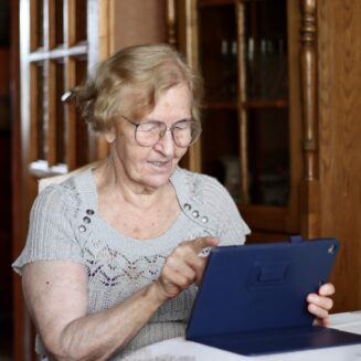 Ältere Frau mit Brille sitzt an einem Tisch und benutzt ein Tablet in einem gemütlichen Raum