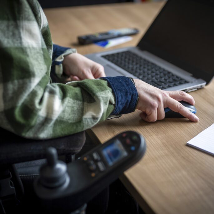 Person im Rollstuhl benutzt einen Computer mit Tastatur und Maus an einem Holztisch
