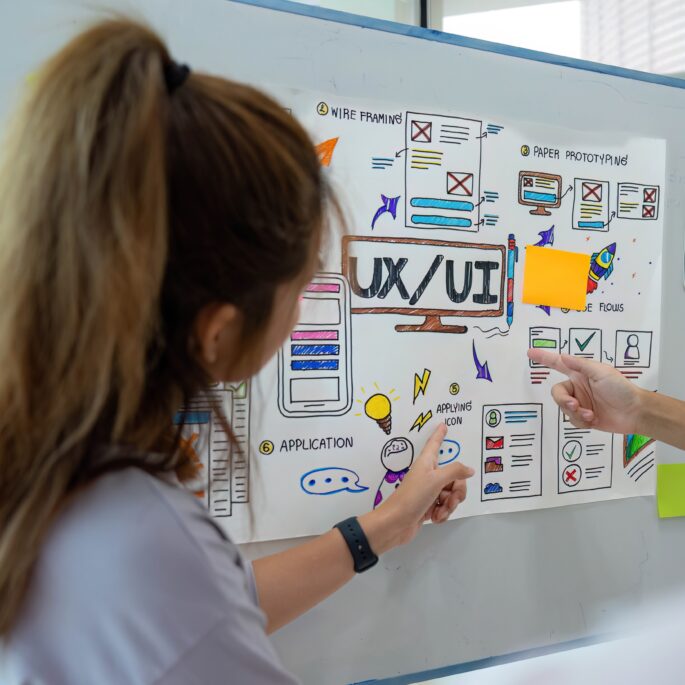 Zwei Personen stehen vor einem Whiteboard mit Skizzen und Notizen zu UX/UI Design, sie zeigen auf die gezeichneten Konzepte