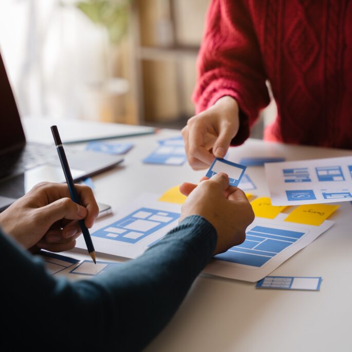 Zwei Personen arbeiten gemeinsam an einem Tisch, sie entwerfen Website-Layouts auf Papier mit Markierungen und Notizen