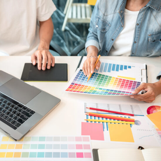 Zwei Personen arbeiten an einem Tisch mit Farbkarten, einem Laptop und verschiedenen Designmaterialien