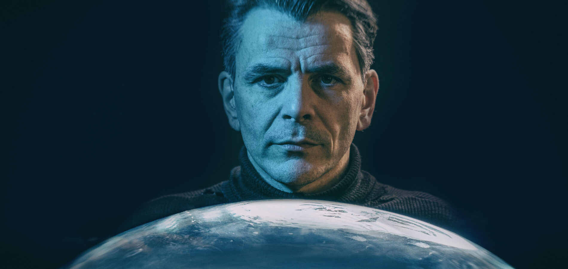 Porträt eines Mannes im blauen Licht, der hinter einer gläsernen Kuppel steht, in der eine Stadt zu sehen ist