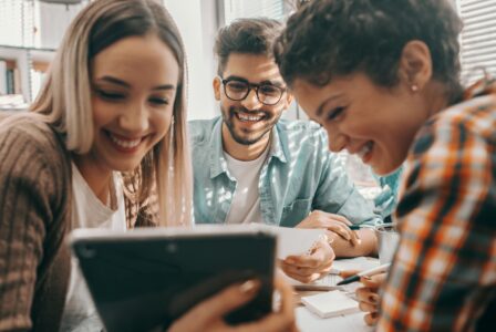 Drei junge Erwachsene sitzen zusammen an einem Tisch und schauen gemeinsam auf ein Tablet, dabei lachen sie und wirken motiviert