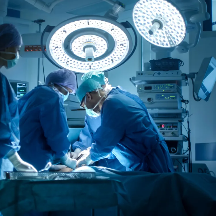 Ärzte und medizinisches Personal führen eine Operation im Krankenhaus unter hellem OP-Licht durch. Im Hintergrund sind medizinische Geräte und Monitore zu sehen.