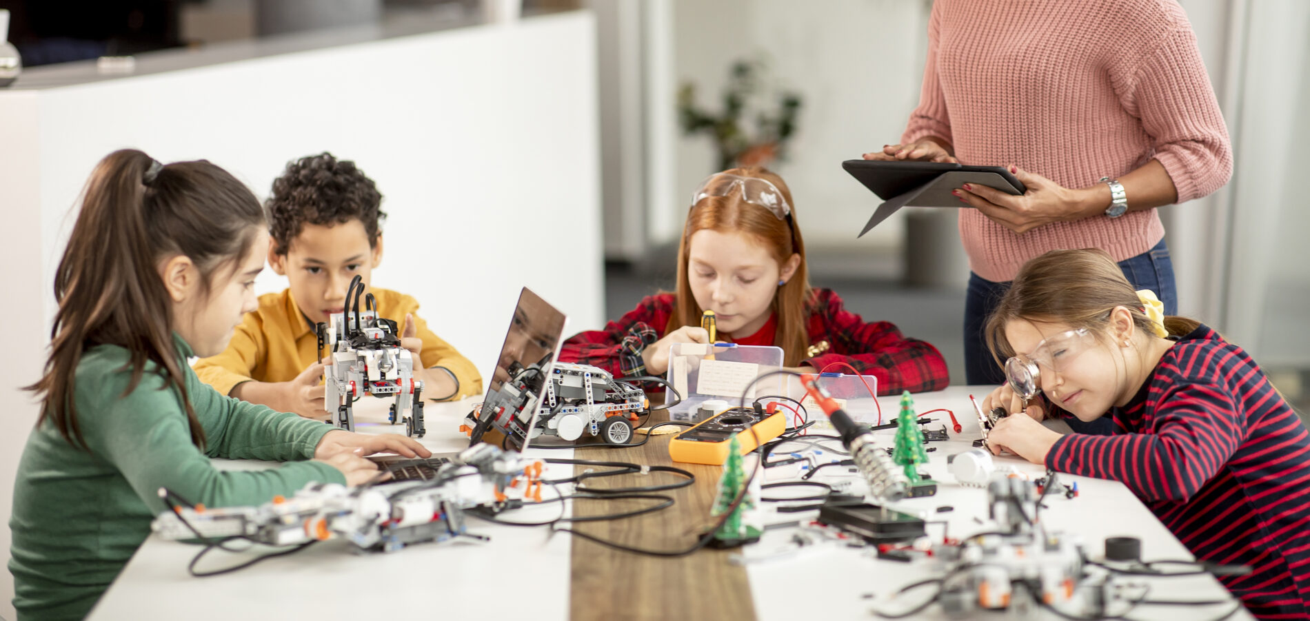 Vier Kinder sitzen um einen Tisch mit kleinen Robotern und einem Laptop, sie arbeiten, eine Frau steht mit einem Tablett daneben und schaut auf die Kinder