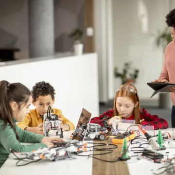 Vier Kinder sitzen um einen Tisch mit kleinen Robotern und einem Laptop, sie arbeiten, eine Frau steht mit einem Tablett daneben und schaut auf die Kinder
